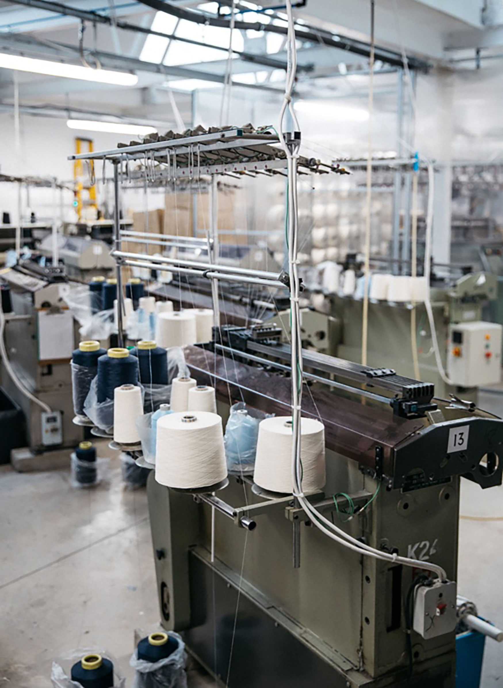 Photograph of the inside of the factory, shows large green textile machines, with white and blue thread rolls in poles, and also other textile machinery. Shows a skylight in the roof