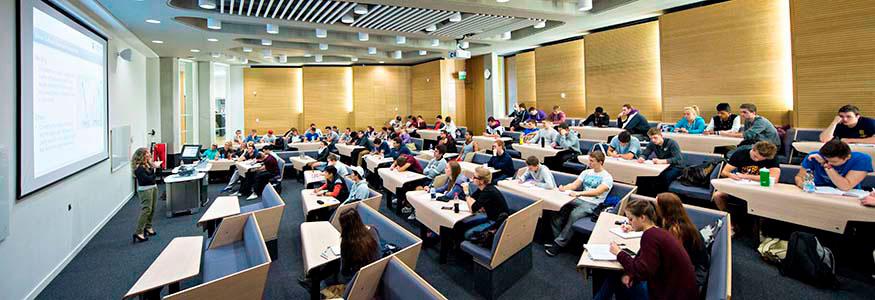 Photo of the conference centre, shows people sitting at small conference desks looking at the front where the lecturer is giving a presentation