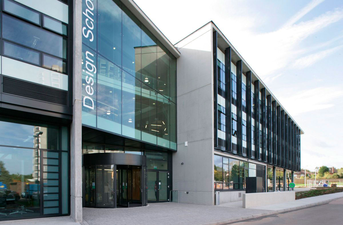 Photo of the Design School, shows a grey and cool green front made out of a lot of glass, which very light grey concrete panels, then the glass revolver door at the front