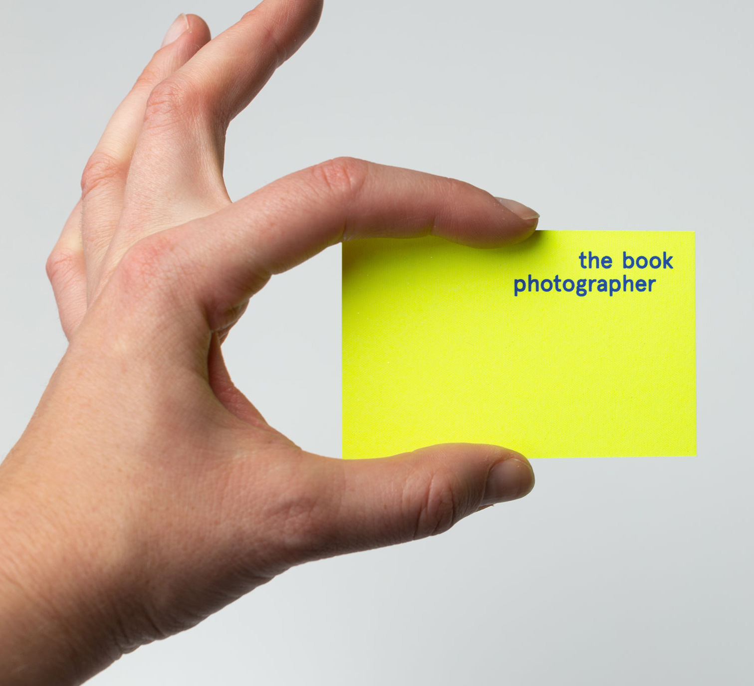 Photograph of a hand on the left, holding a bright fluorescent yellow business card between the index finder and thumb, with the words ‘The Book Photographer’ written on it in bold blue letters