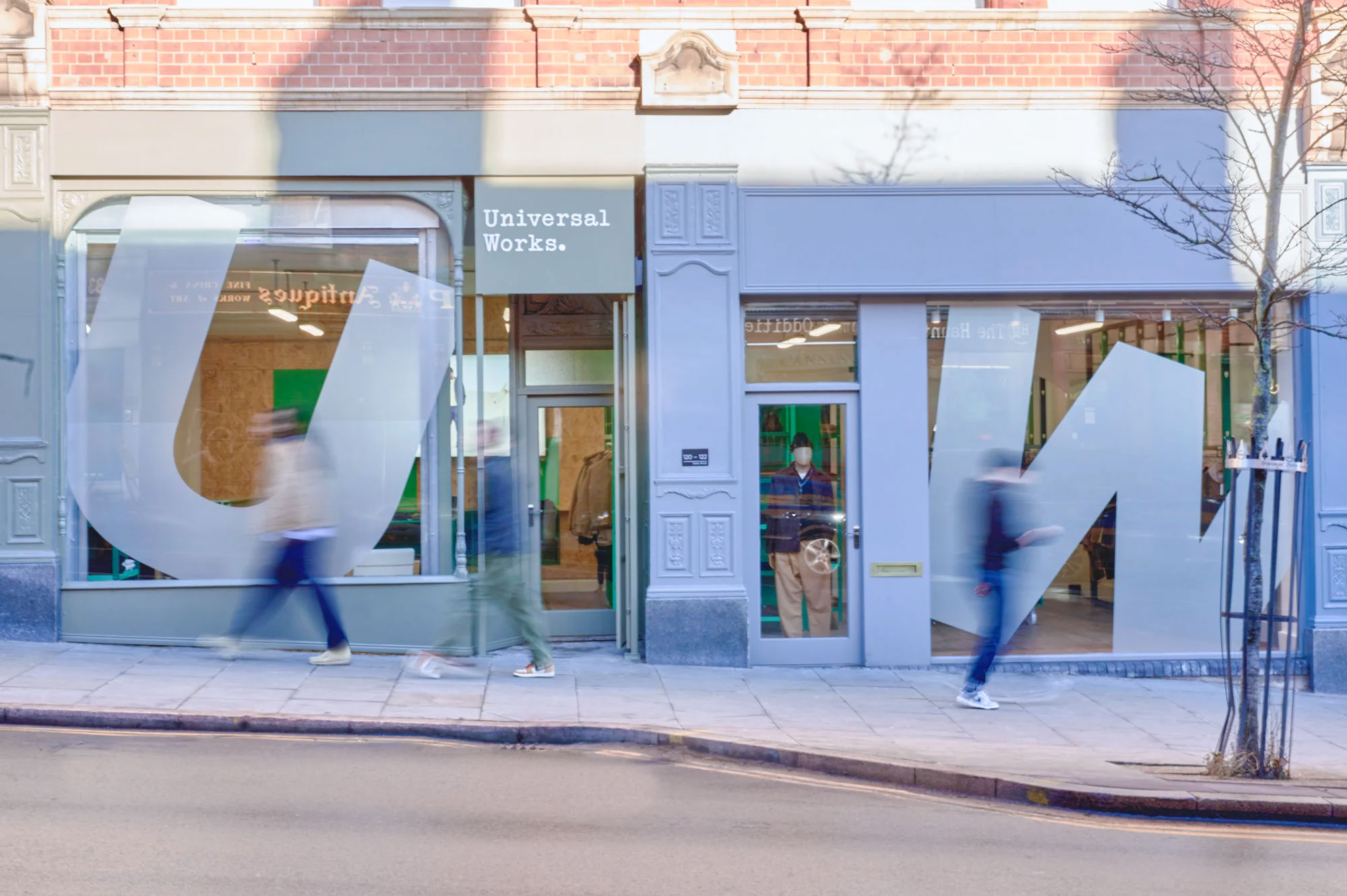 Photograph of the shop front, shows the grey painted front, 1 room on the left with a large window, same on the right. Then people walking by blurry. With a road going bye below