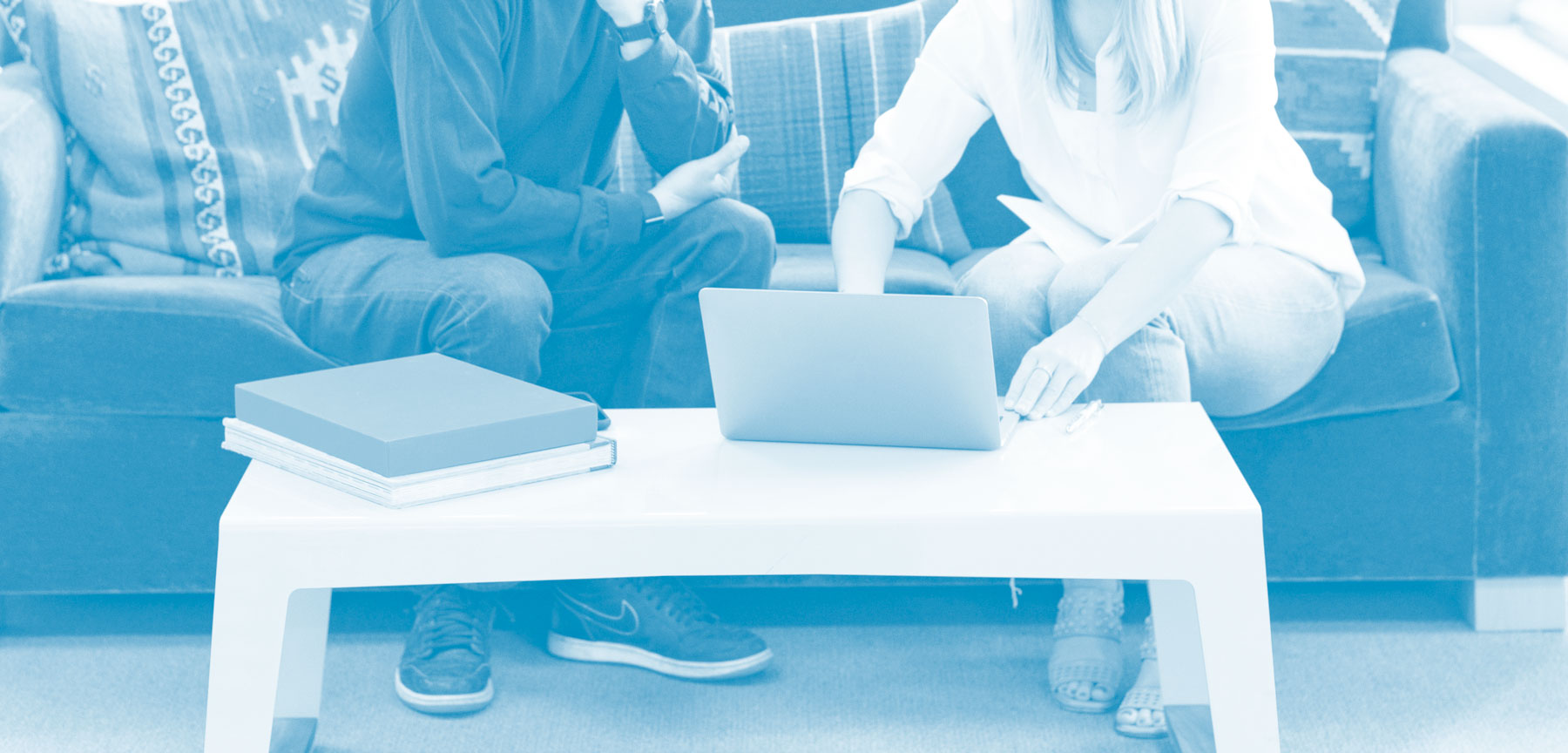 Photo of a large sofa, with a man sitting on the left and a woman on the right, the man is asking the woman questions (user testing), there is a white table in front, with a laptop on, and books on the left