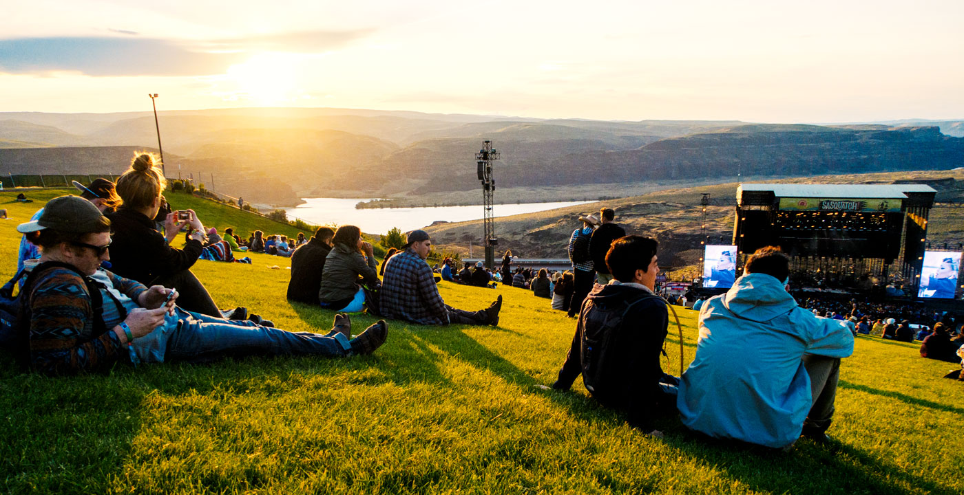 Colour grade added (moody and darker effect) photo of festival people sitting on a hill, looking down the hill to the main stage. Light sunset and mountains in the background