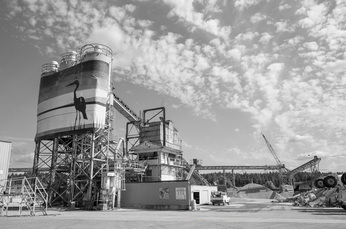 Uncorrected colour photograph of an industrial site showing yellow stairs and large chemical tanks on the left, with a haulage container and truck on the right, with the sky and trees in the background. This is 1st black and white example