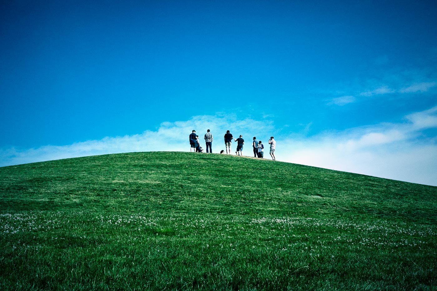 Original photo of a green hill in the front and blue sky in the background with a group of people standing on the top of this hill. Vintage colour grading applied