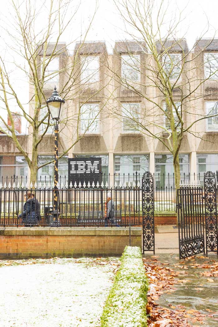 Shows New Walk (a lane) going across the middle of the photo, with the IBM office behind it which is a light grey concrete building. Show an iron gate at the front and people walking behind it down New Walk