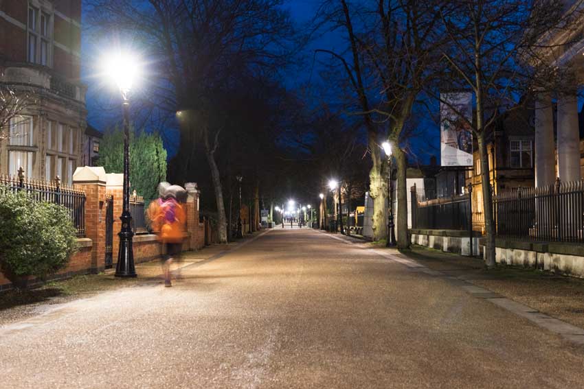 Photography at night time outside Leicester Museums and Galleries, shows a bright street light on the left, and a blurred woman jogger with an orange top on running past the museum which is on the right