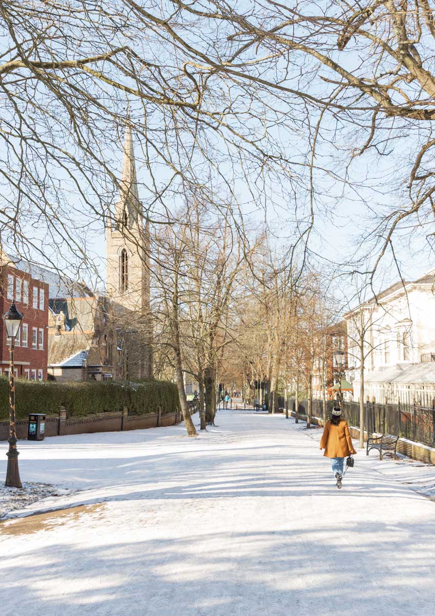 Shows New Walk (a lane) going down, an old lamppost on the left, with a large church also on the left. Then shows a woman walking down New Walk with a bright yellow mustard coat on. The photograph is in the winter and shows the snow on the ground