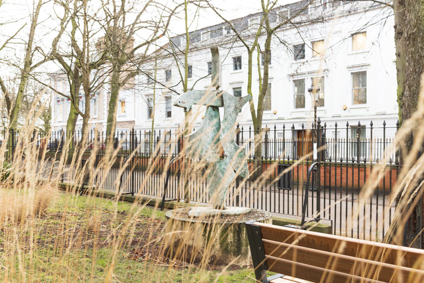 Shows The Clicker sculpture by John Atkin on New Walk, with iron railings and offices behind it, then straw like yellow thin plants waving in the breeze, in front of the sculpture. The sculpture is a green marbled abstract figure of a person